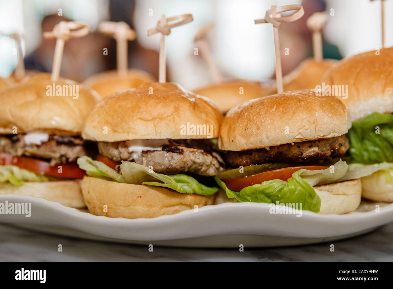 Burgers with beef patty, fresh tomato, lettuce, fried onion and melted cheese held with a wooden skewer, served on a white ceramic plate. Stock Photo