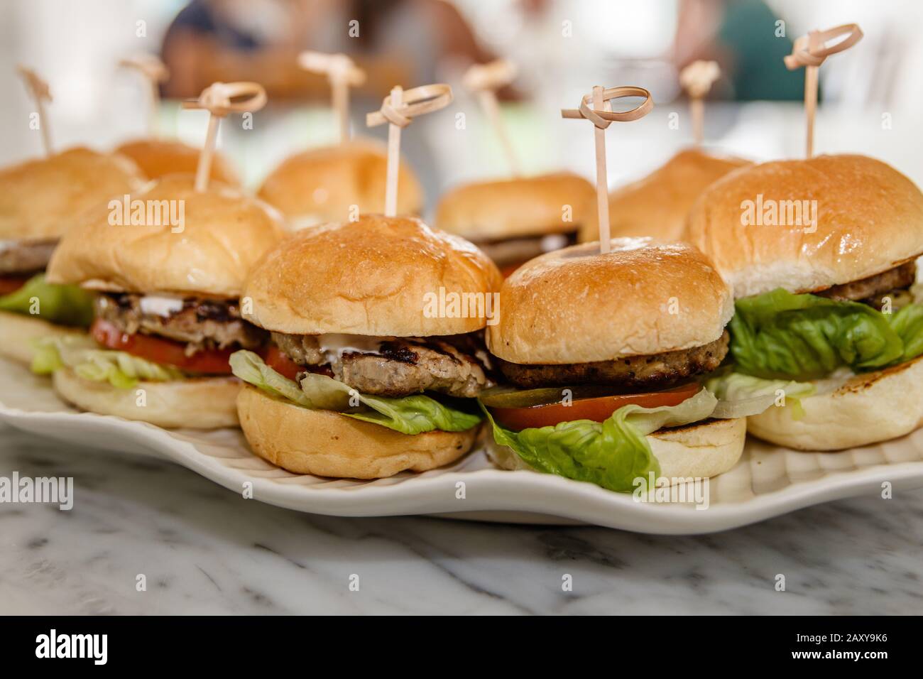 Burgers with beef patty, fresh tomato, lettuce, fried onion and melted cheese held with a wooden skewer, served on a white ceramic plate. Stock Photo