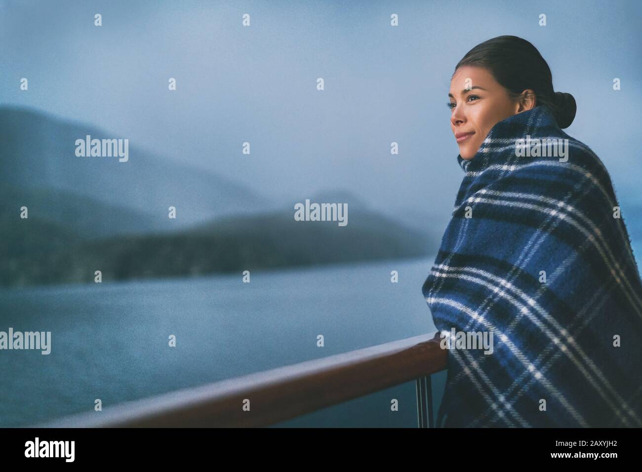 Cruise ship passenger on Alaska travel vacation enjoying scenery at dusk on suite balcony deck with wool throw in cold weather. Asian tourist woman relaxing on summer holiday cruising adventure. Stock Photo