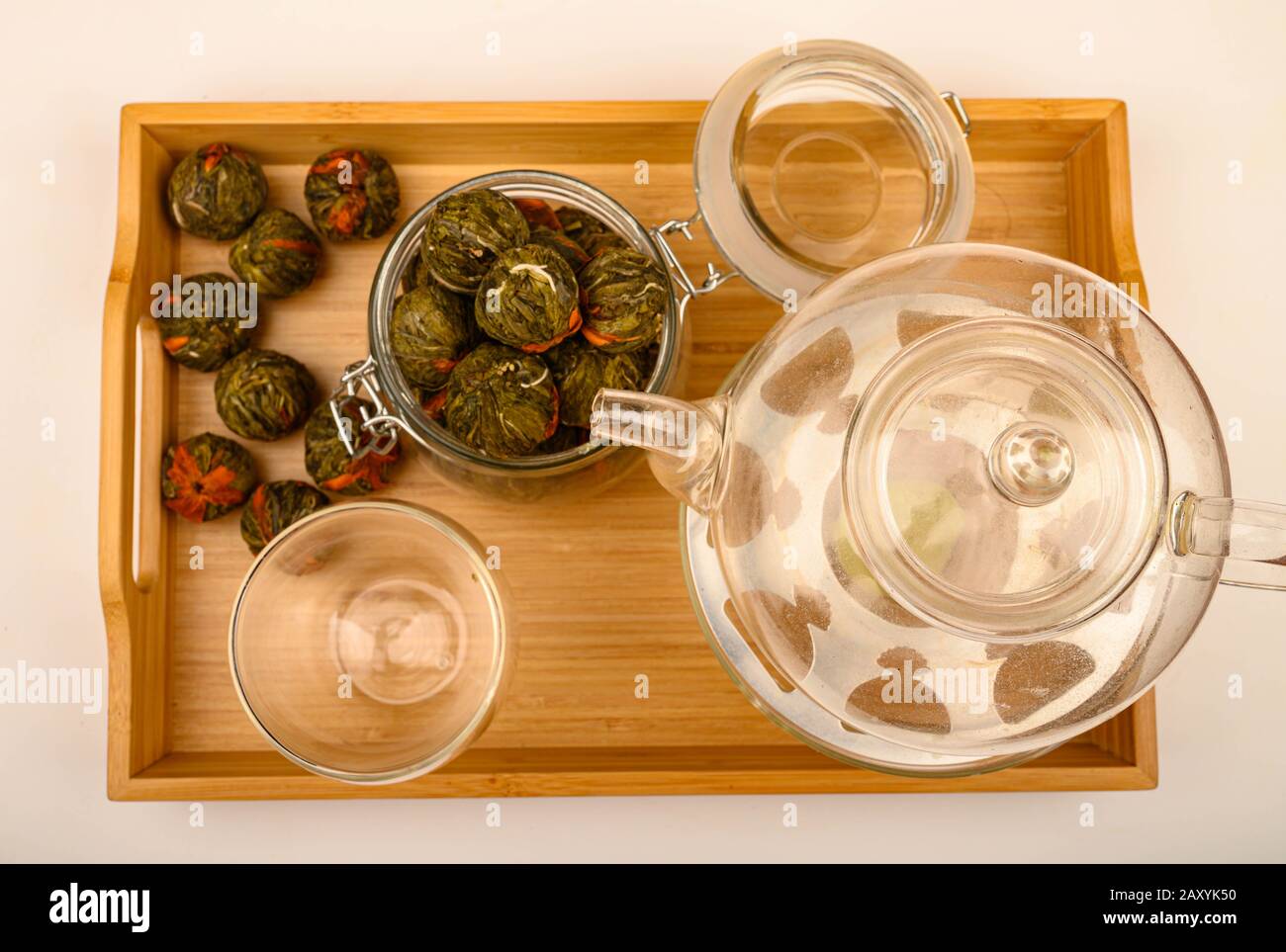 Flower tea balls in a glass jar, a glass teapot and a glass on a wooden tray on a white background. Close up Stock Photo