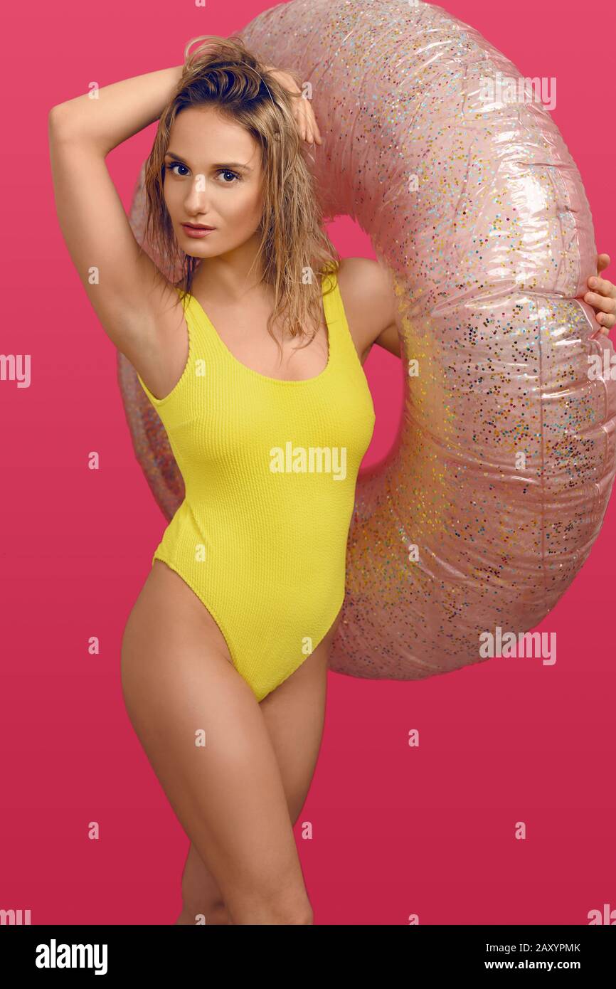 Sexy slender young woman in yellow swimsuit holding a large inflatable tube over her arm as she smiles at the camera with hand on hip over a red backg Stock Photo