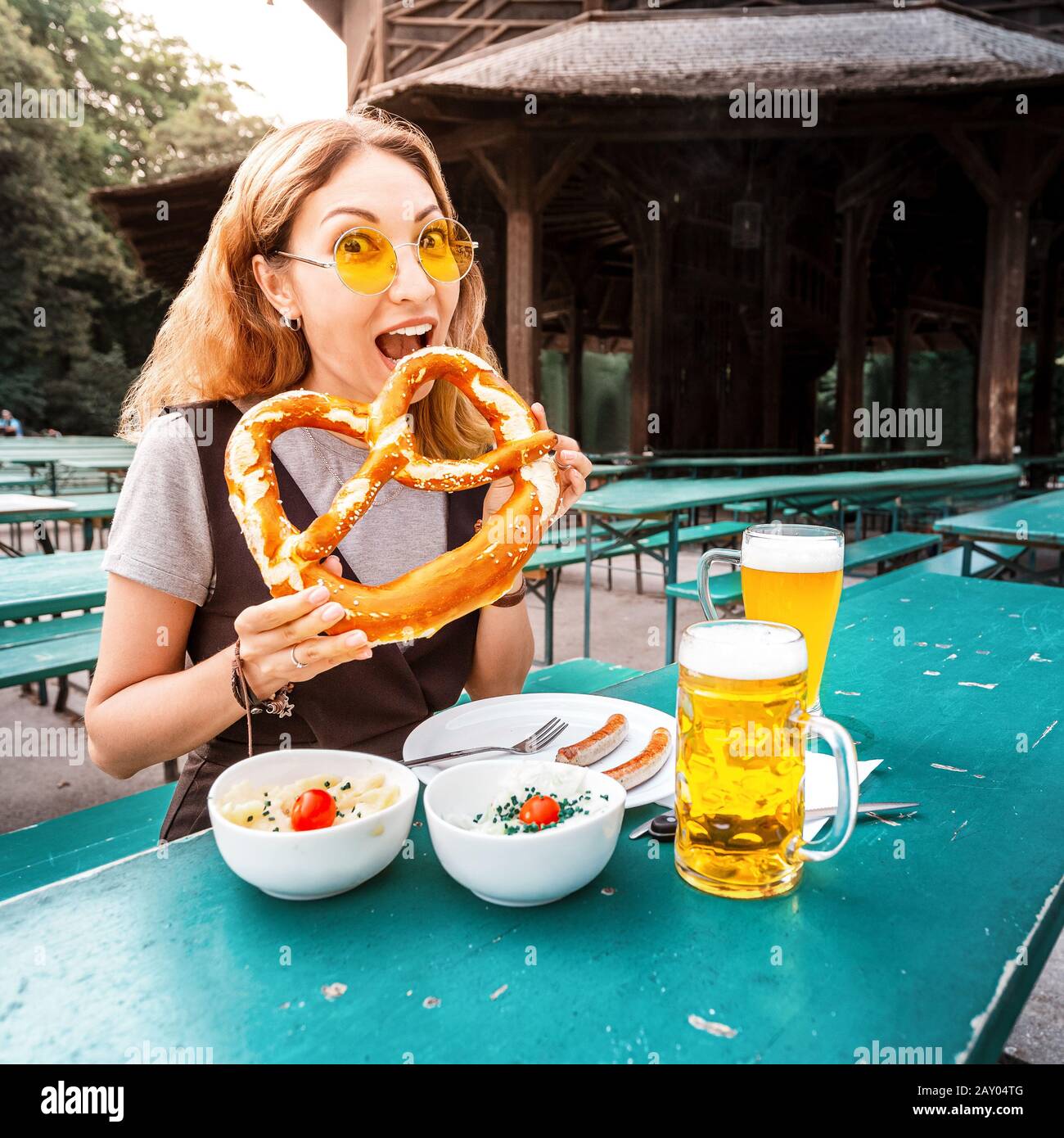 Asian girl eating traditional Pretzel and drinking fresh Bavarian beer in beer garden in Munich. The concept of traditional food festival and tourism Stock Photo