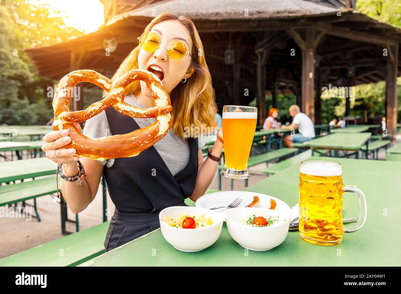 Asian girl eating traditional Pretzel and drinking fresh Bavarian beer in beer garden in Munich. The concept of traditional food festival and tourism Stock Photo