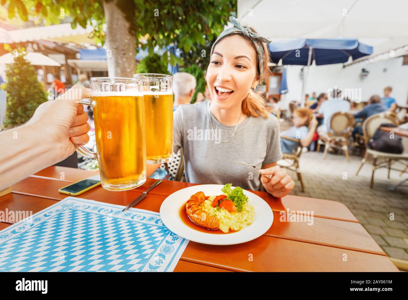 Happy Asian girl clinks a mug of Bavarian beer in Biergarten. Traditional German snack with sausage and potato salad Stock Photo