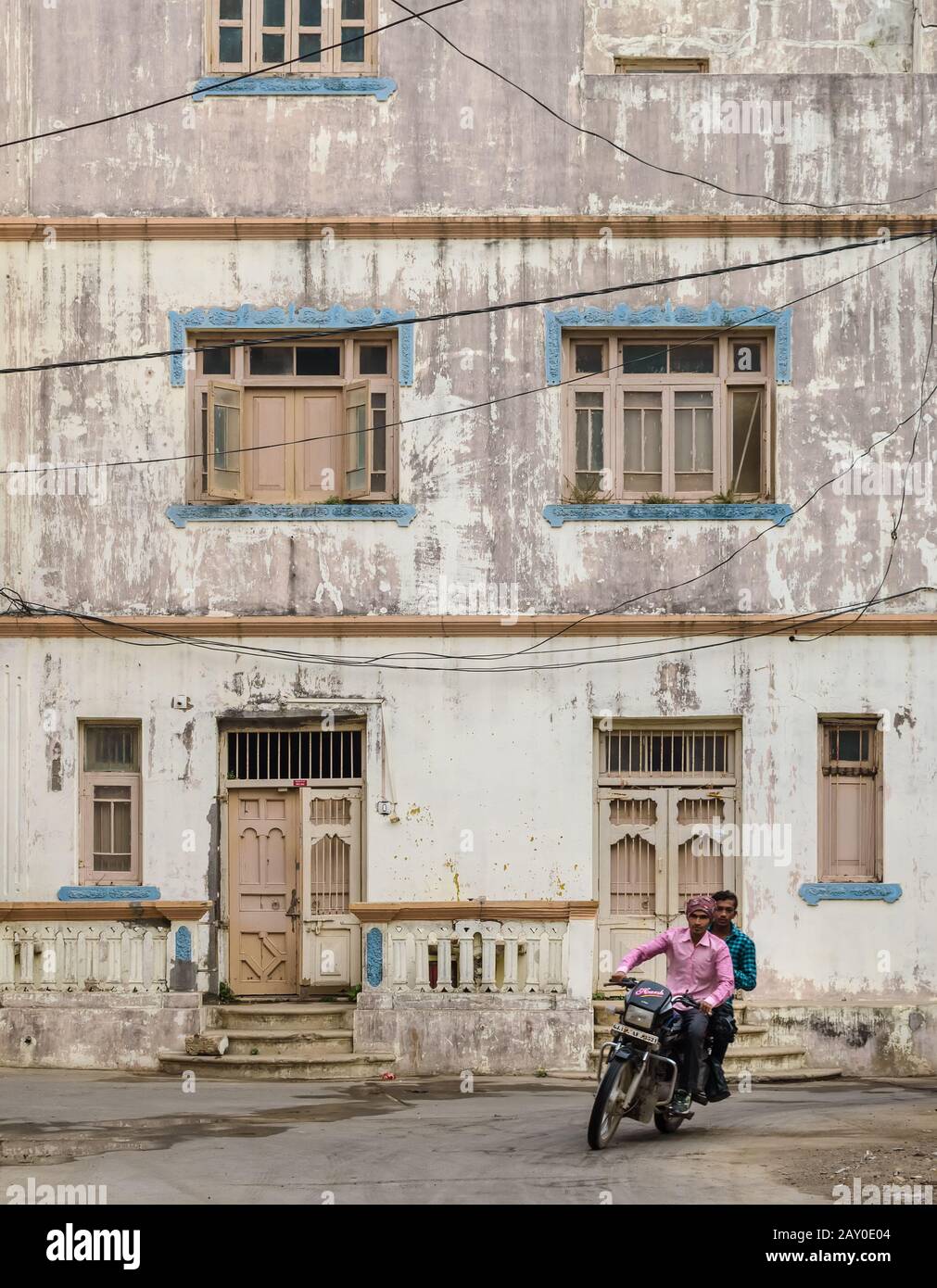 Diu, India - December 2018: Two men bike down a road past an old weathered residential building with quaint windows and wooden doors. Stock Photo