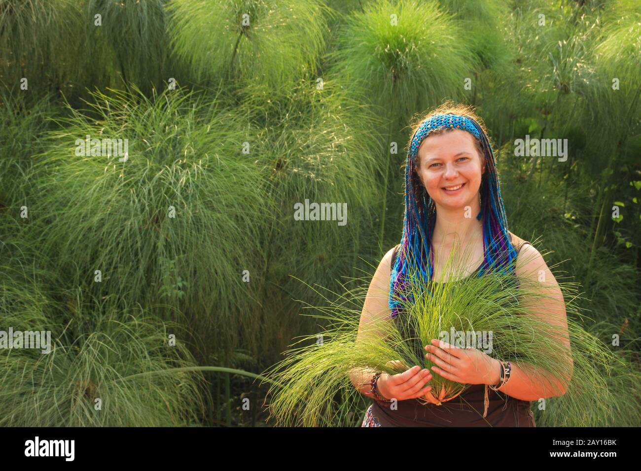 A young Caucasian woman with blue pigtails stands against a field with papyrus and holds papyrus flowers in her hands. Stock Photo