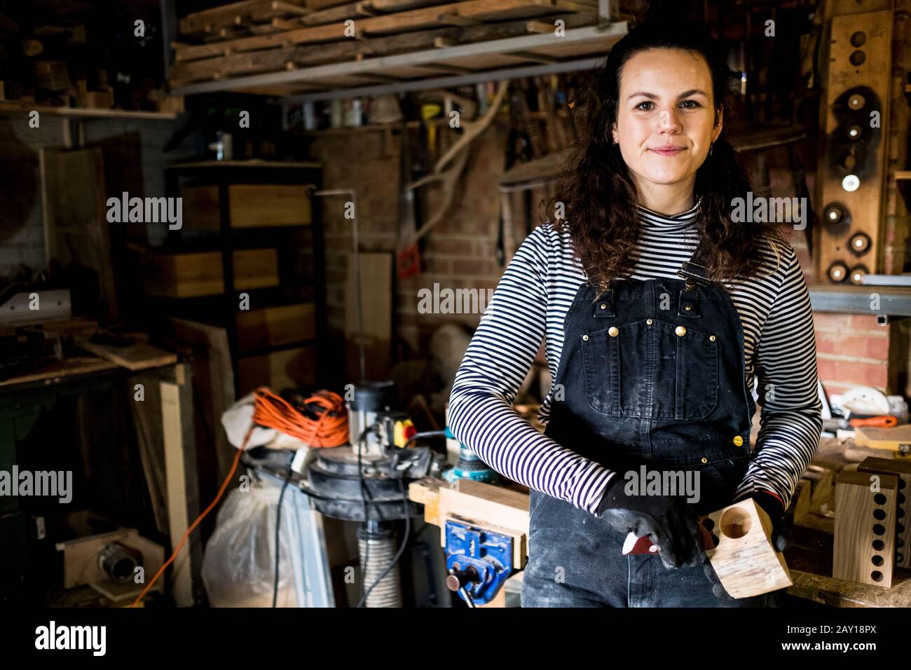 Woman with long brown hair wearing dungarees standing in wood workshop ...