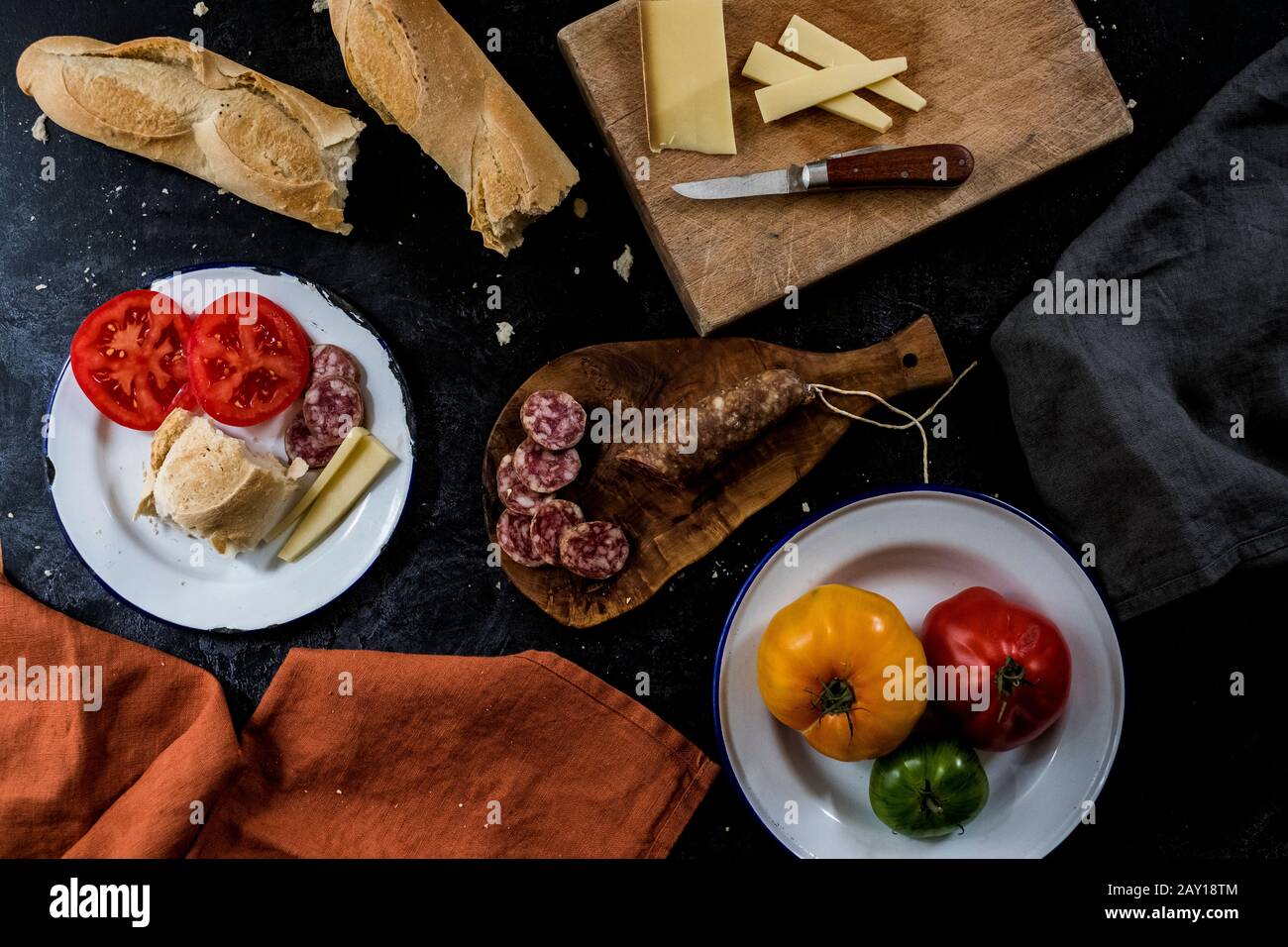High angle close up of a selection of cheeses, tomatoes, salami and French baguette on white enamel plates on black background. Stock Photo