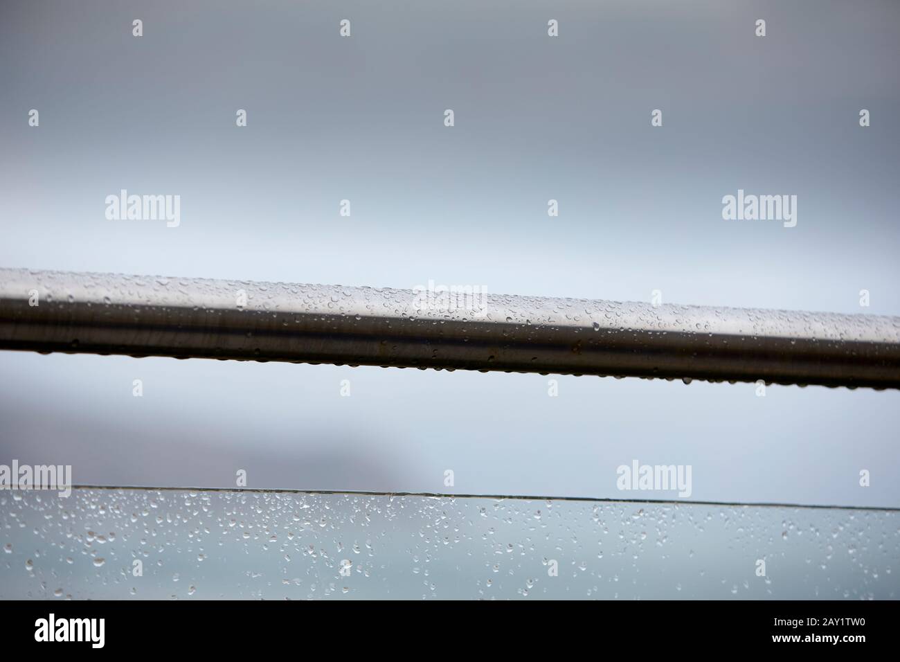 Stainless steel handrail and glass safety panel balustrade close up with raindrops against soft focus blurred overcast grey sky, north wales coast uk Stock Photo