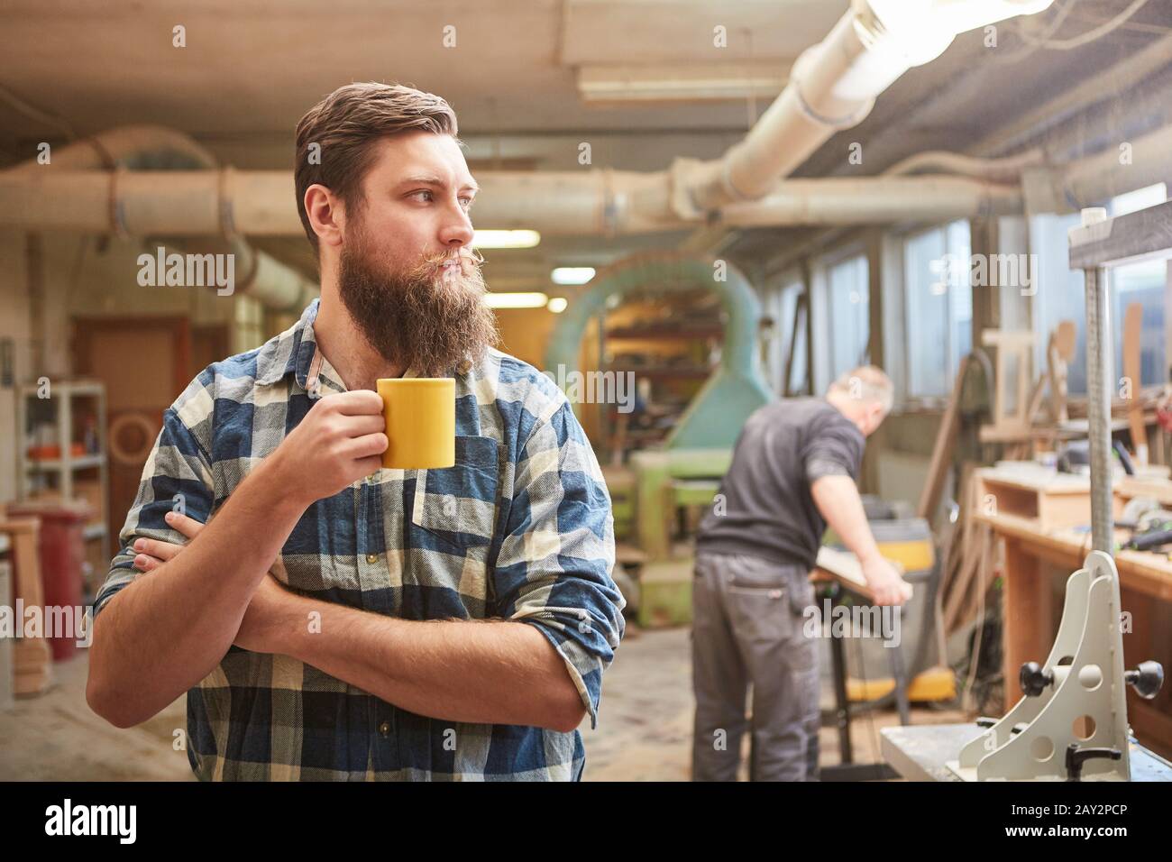 Carpenter with a beard drinks a cup of coffee in the joinery during a break Stock Photo