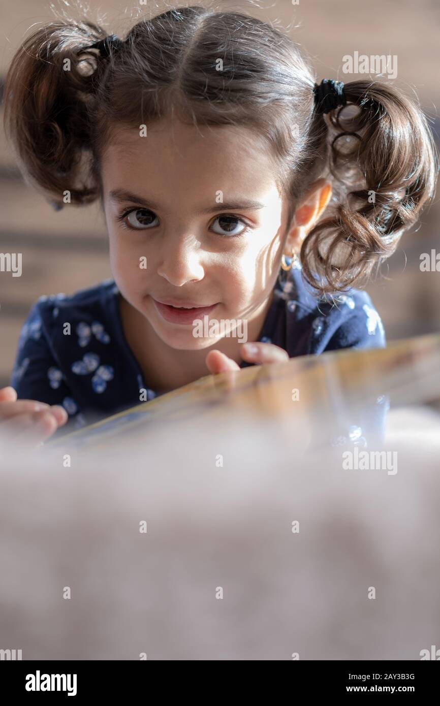 Middle Eastern young cute girl lies on a light bed in the light of sunlight. Persian swarthy girl on the bed. Middle Eastern kids on the bed. Stock Photo