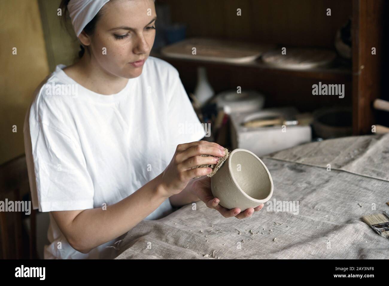 Beautiful woman in a creative workshop sits at a table and wipes a clay bowl Stock Photo