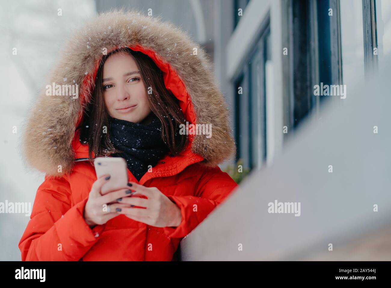 Photo of attractive woman holds modern modern gadget, wears red jacket with hoody, checks email box, connected to wireless internet, stands outdoor, e Stock Photo