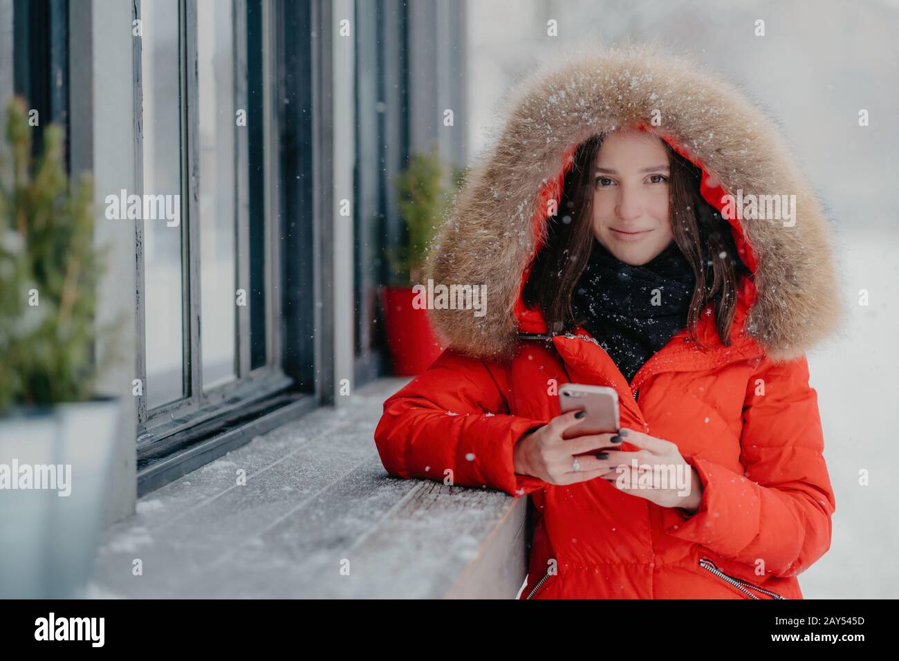 Smiling young European female in red coat, has black manicure, uses modern electronic gadget for communication in internet with friends, likes winter Stock Photo