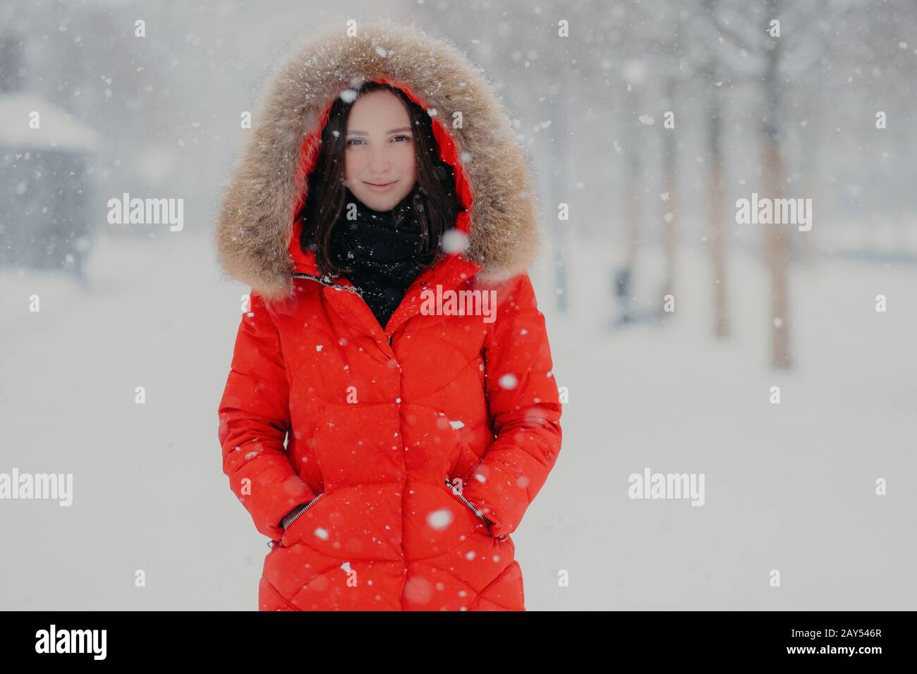 Good looking young woman has appealing look, wears warm red jacket, keeps hands in pockets, has outdoor stroll during winter time and snowfall, poses Stock Photo