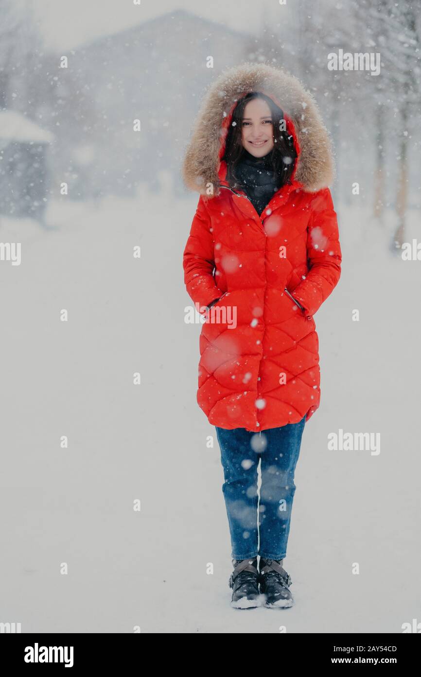 Full length vertical shot of pretty woman wears red winter jacket, jeans and boots, keeps hands in pockets, stands on street during winter time during Stock Photo