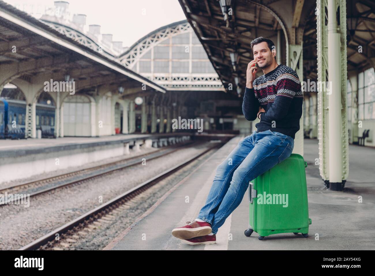 Everyday lifestyle concept. Handsome man wears sweater and jeans, poses at rail station platform, leans at bag, has telephone conversation, focused in Stock Photo