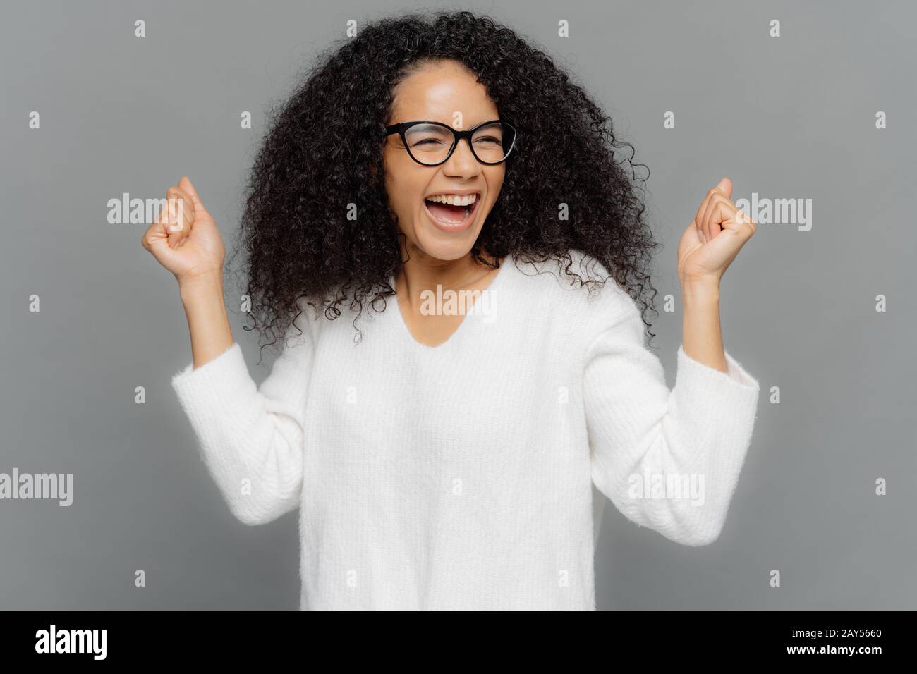 Studio shot of triumphing dark skinned woman clenches fists, smiles happily, celebrates success, achieves goal, smiles broadly, dressed casually, isol Stock Photo