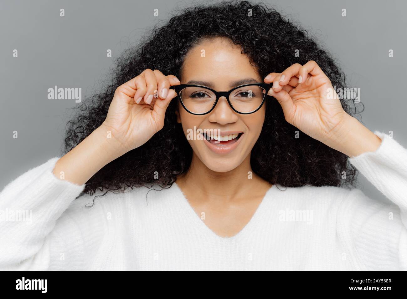 Close up shot of pretty dark skinned female has minimal makeup, keeps hands on frame of transparent glasses, dressed casually, enjoys spare time and g Stock Photo