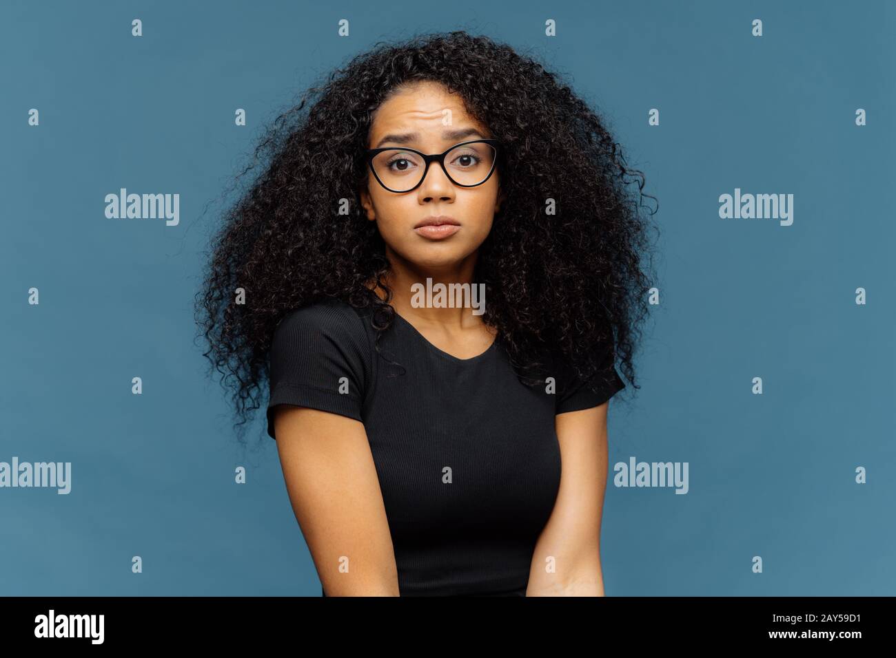 Photo of indignant puzzled Afro American woman looks surprisingly at camera, wears casual black t shirt and spectacles, poses against blue background. Stock Photo