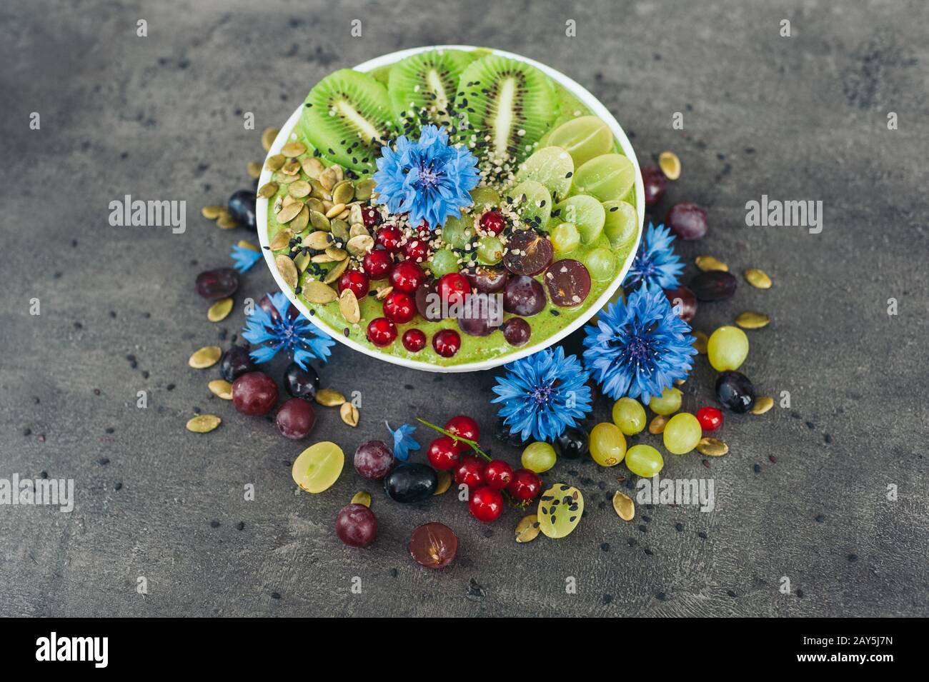 Bowl with fresh kiwi salad, seeds, red current, green grape and blue cornflowers around. Vegeterian dish of ripe fruit. Top view. Blank space Stock Photo