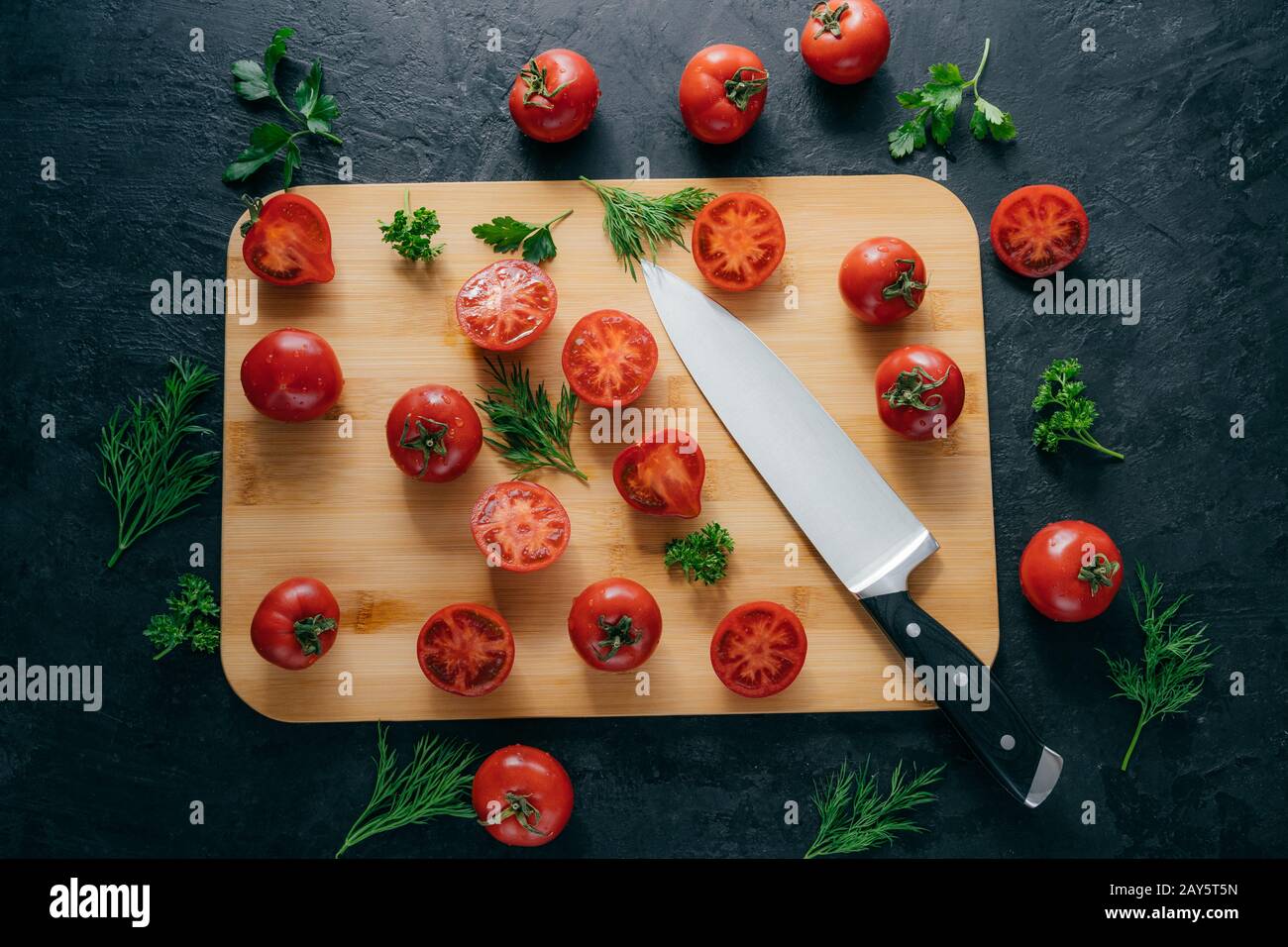 Top view of red sliced tomatoes on wooden chopping board. Sharp knife near. Green parsley and dill. Dark background. Preparing fresh vegetable salad Stock Photo