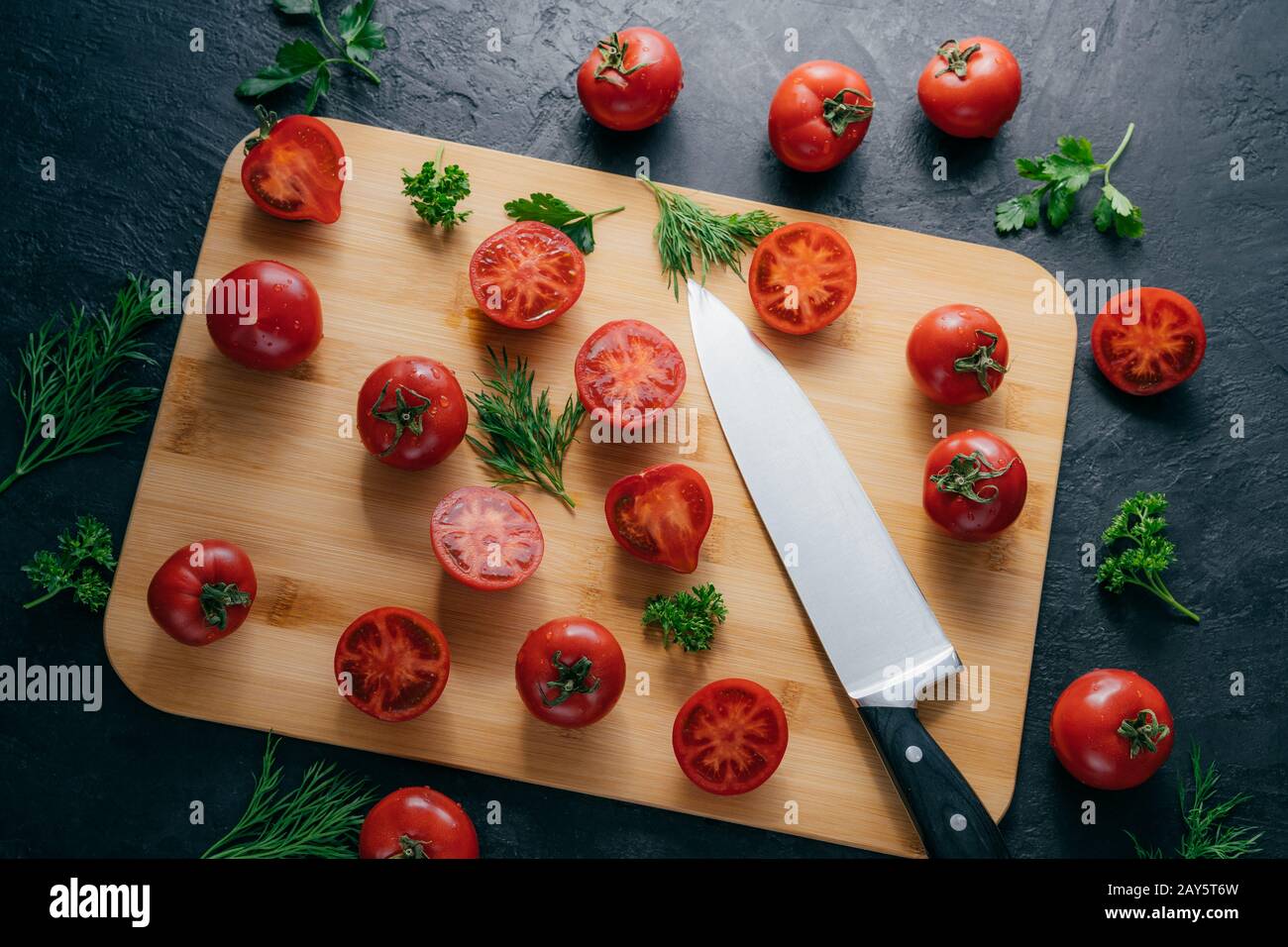 Tasty ripe tomatoes sliced on chopping board with greenery, sharp knife near, isolated over dark background. Kitchenware. Top view. Stock Photo