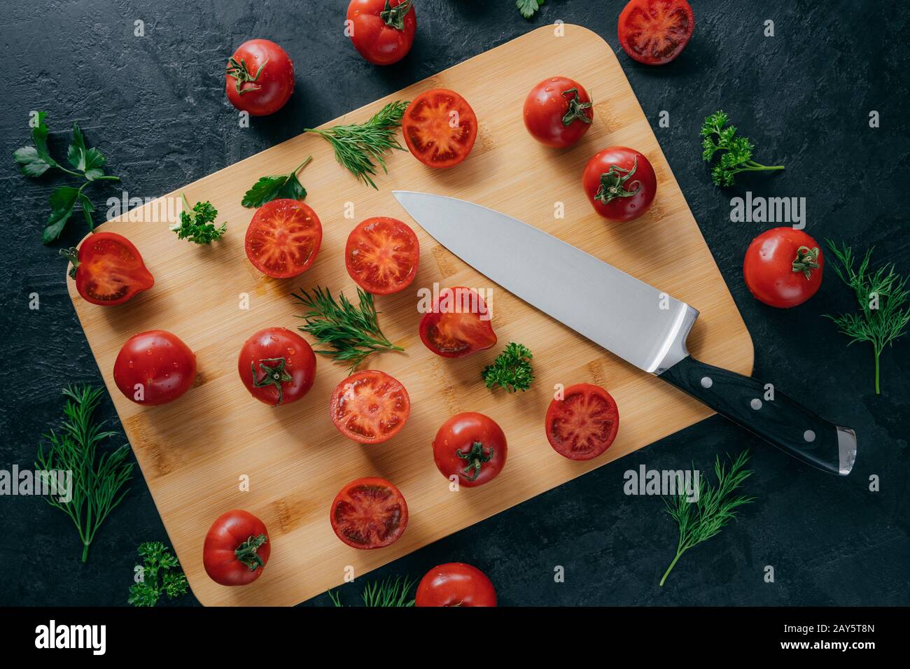 Top view of fresh small red tomatoes sliced on wooden kitchen board with knife. Green parsley and dill near. Vegetables and vitamins Stock Photo