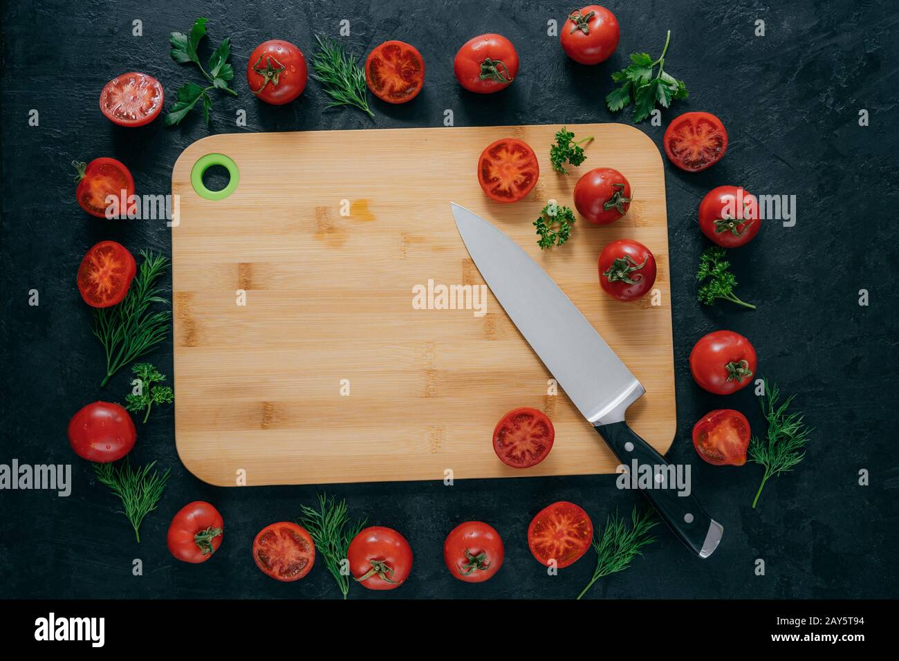 Tomatoes frame around wooden cutting board. Ripe vegetables and slice, green parsley and dill near kitchen board and knife. Food art Stock Photo