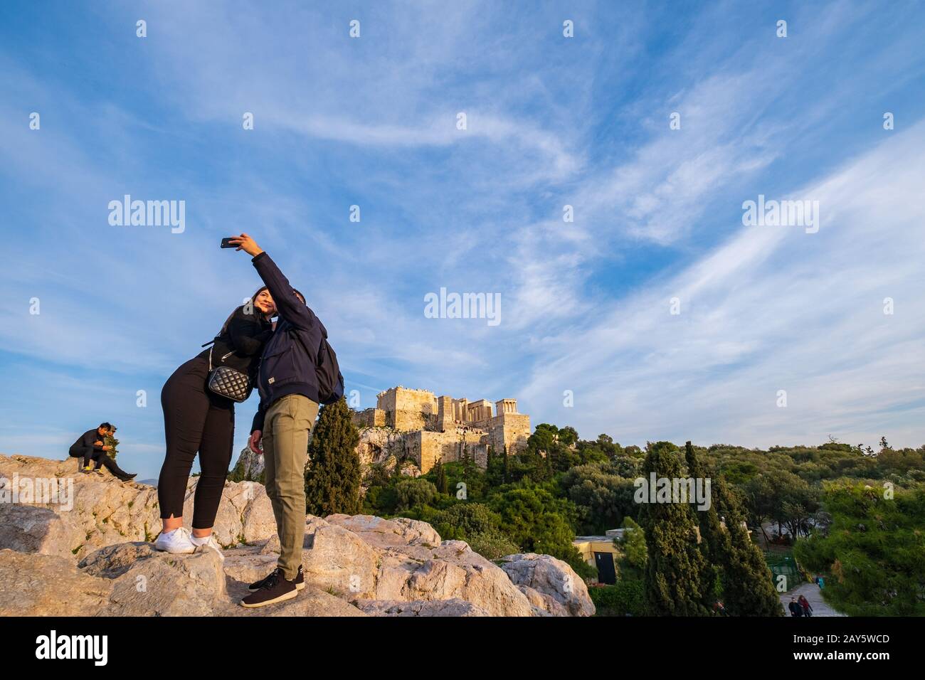 Athens, Greece - January 25, 2020: A couple are taking a selfie on a hill across the Acropolis rock with the Acropolis in the background on a sunny af Stock Photo