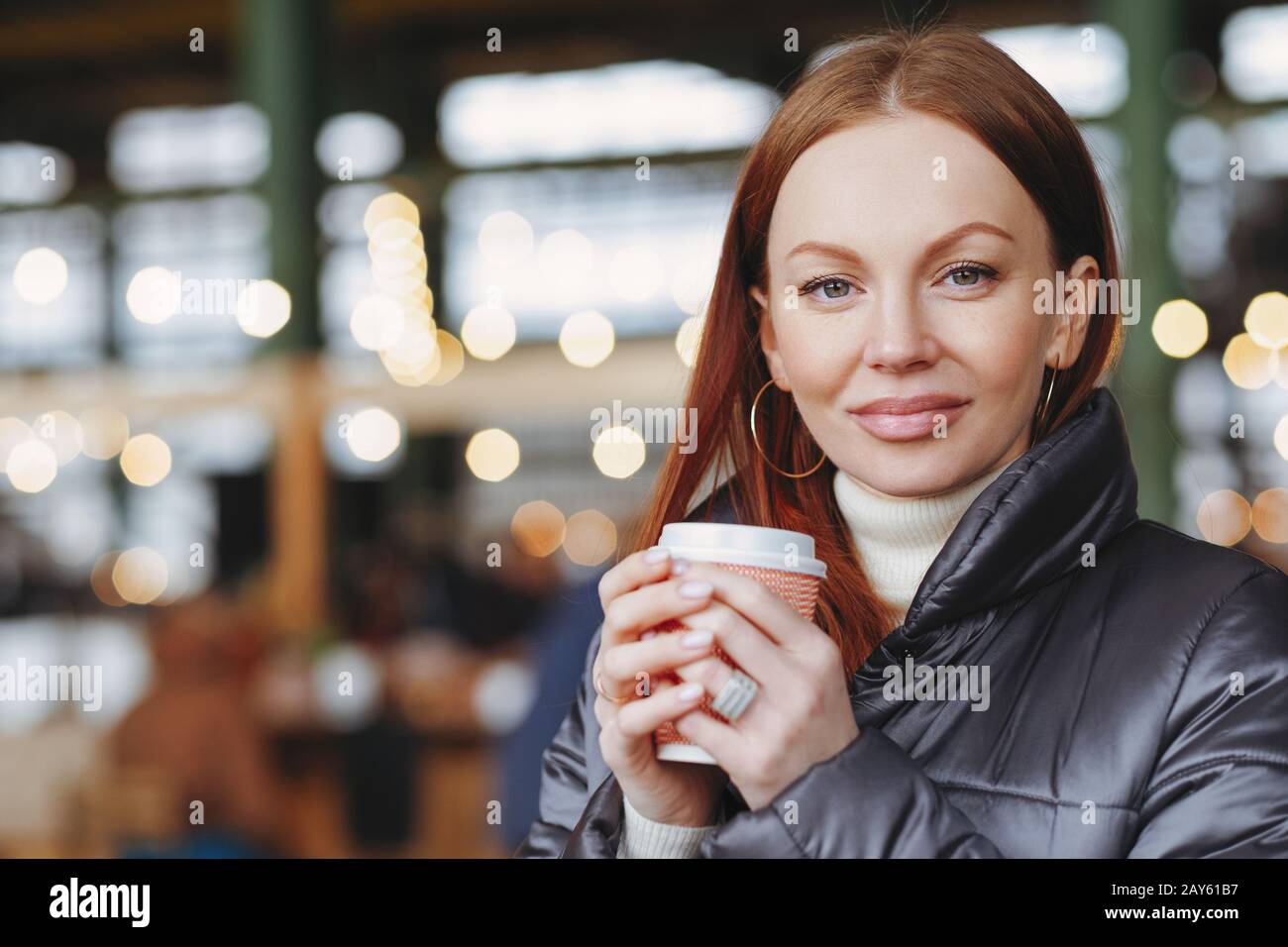 Outside shot of satisfied attractive female model with healthy skin, brown hair, holds takeaway coffee, enjoys good drink, dressed in jacket, waits fo Stock Photo