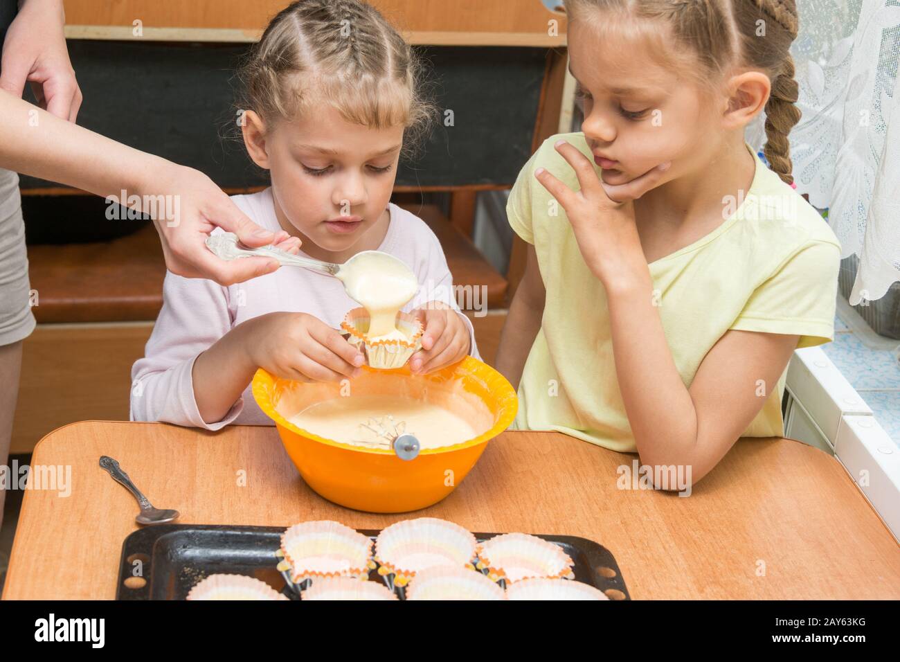Mom helps subsidiaries to pour batter into molds for baking cakes Stock Photo