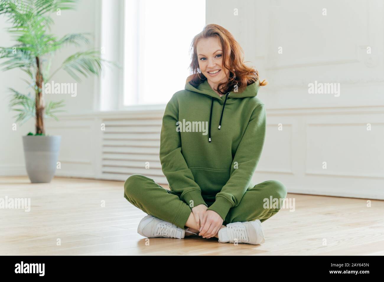 Happy ginger female sport trainer sits crossed legs on floor, wears green hoody and trousers, white trainers, smiles gladfully, poses on floor in empt Stock Photo