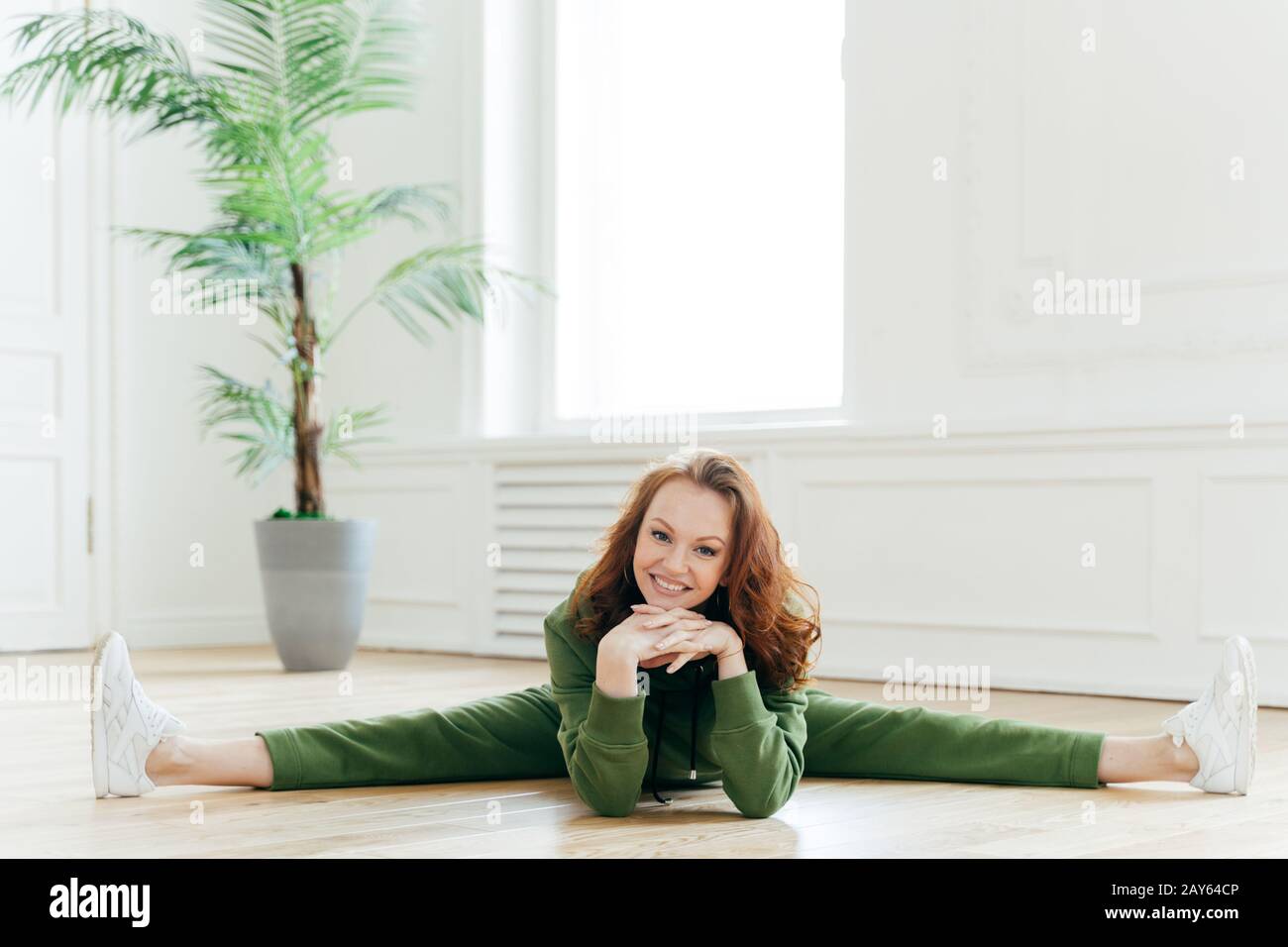 Fitness woman demonstrates nice flexibility, does gymnastics exercises, shows leg split, keeps hands under chin, smiles broadly, poses on floor in spa Stock Photo