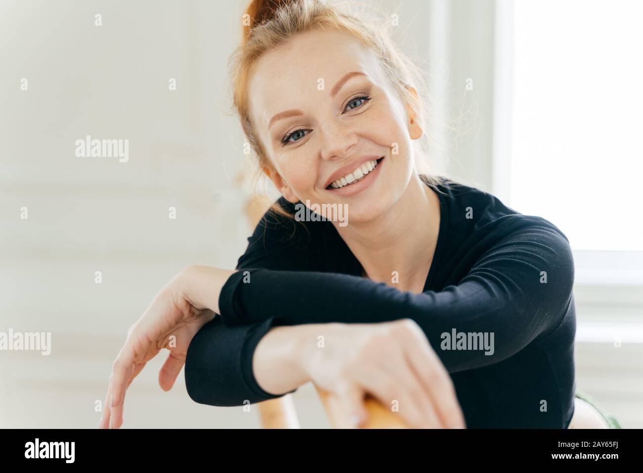 Carefree good looking ginger young female ballerina leans at ballet barre, looks gladfully at camera, practices different dancing positions, has tooth Stock Photo