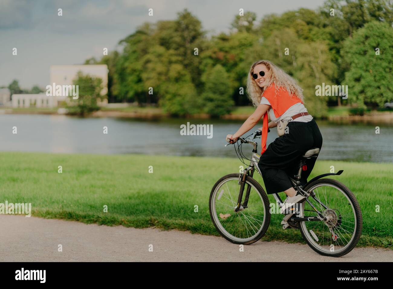 Photo of cheerful woman dressed casually, rides bicycle, looks aside, has happy expression, wears shades, poses near river, green lawn and trees, some Stock Photo