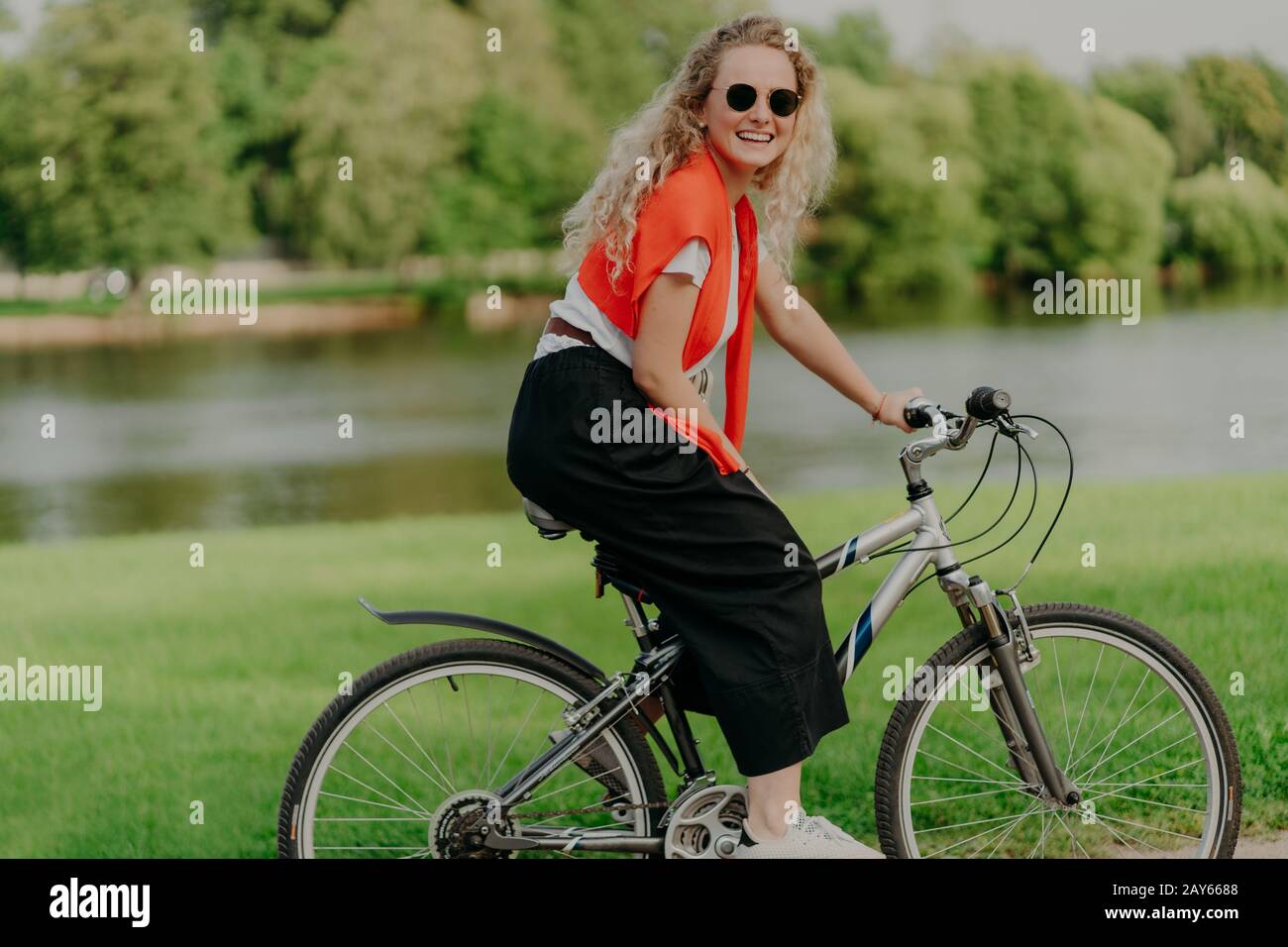 Pleasant looking satisfied curly haired woman spends free time in open air, rides bicycle, spends leisure alone, enjoys freshness of nature, looks at Stock Photo