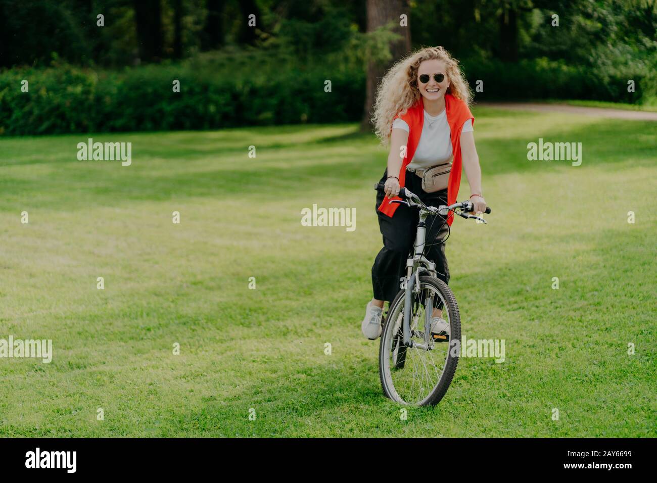 Outdoor image of pretty cheerful young woman rides bike, wears sunglasses, casual wear, poses on green lawn, spends free time in park, bikes in beauti Stock Photo