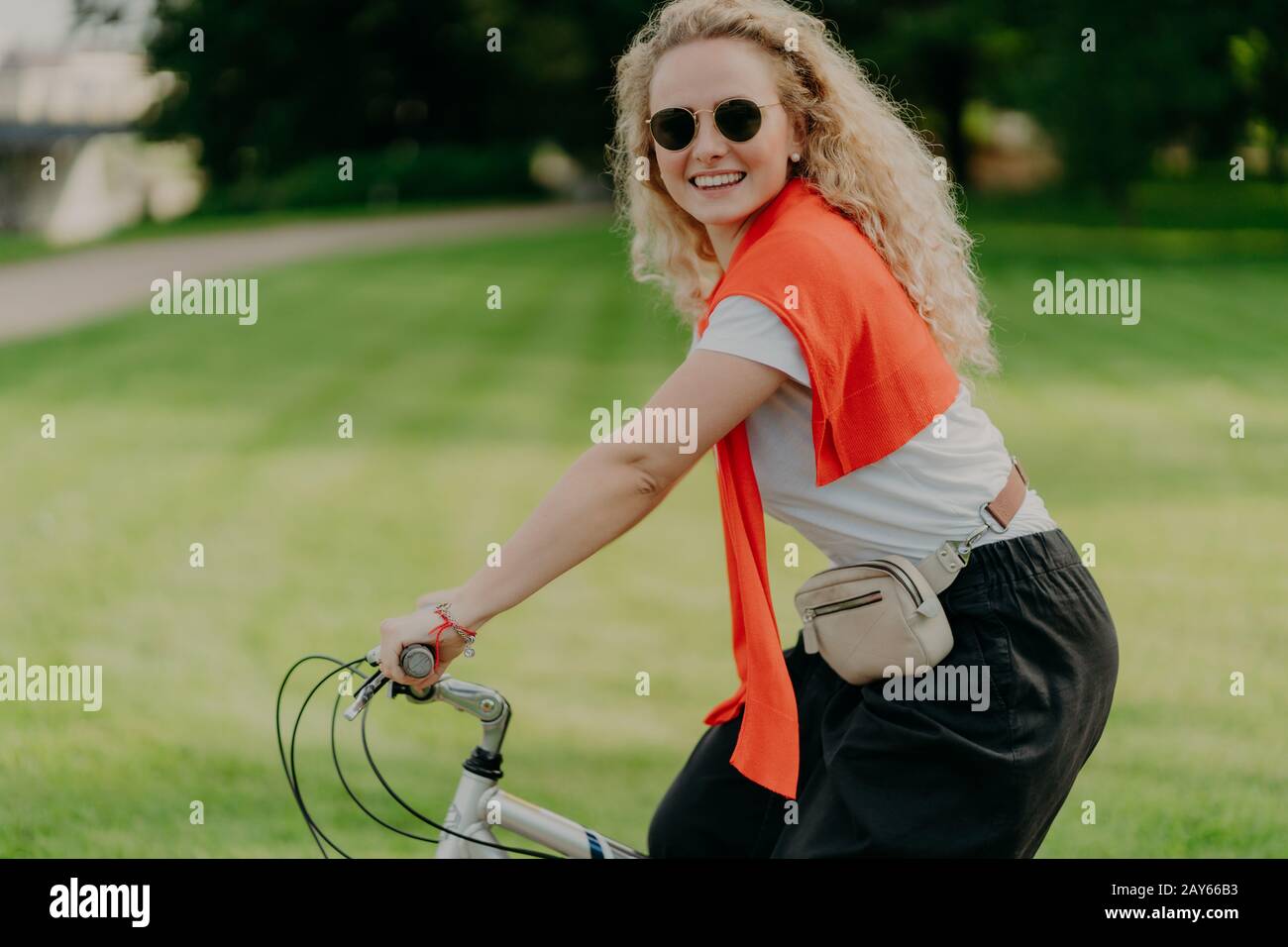 Horizontal shot of happy curly woman rides bike, keeps hands on handlebars, looks active and refreshed, dressed casually, wears shades, poses against Stock Photo