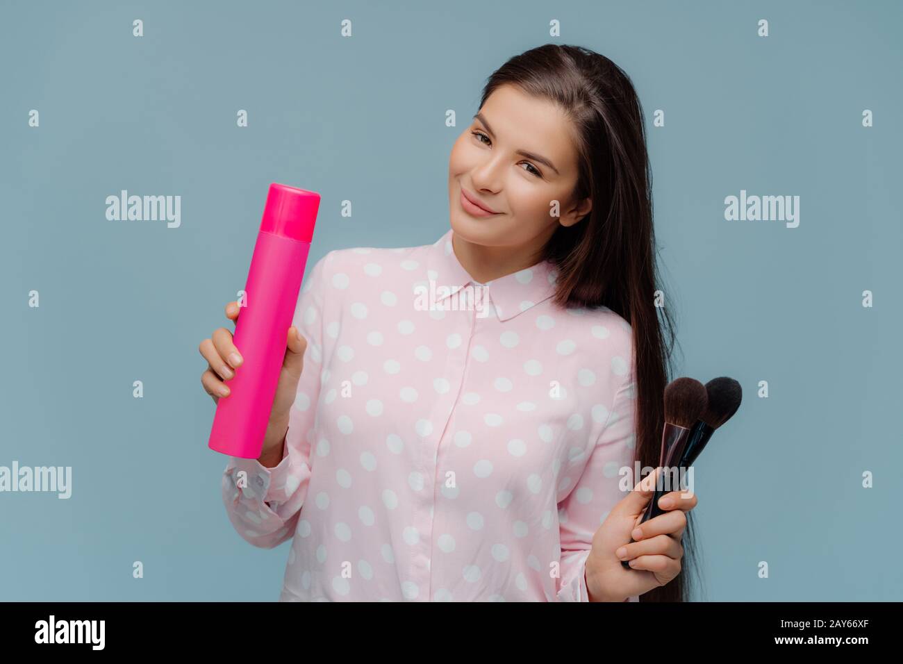 Satisfied long haired brunette female uses hairspray for making stylish hairdo, cosmetic brushes for applying powder on face, prepares for special occ Stock Photo