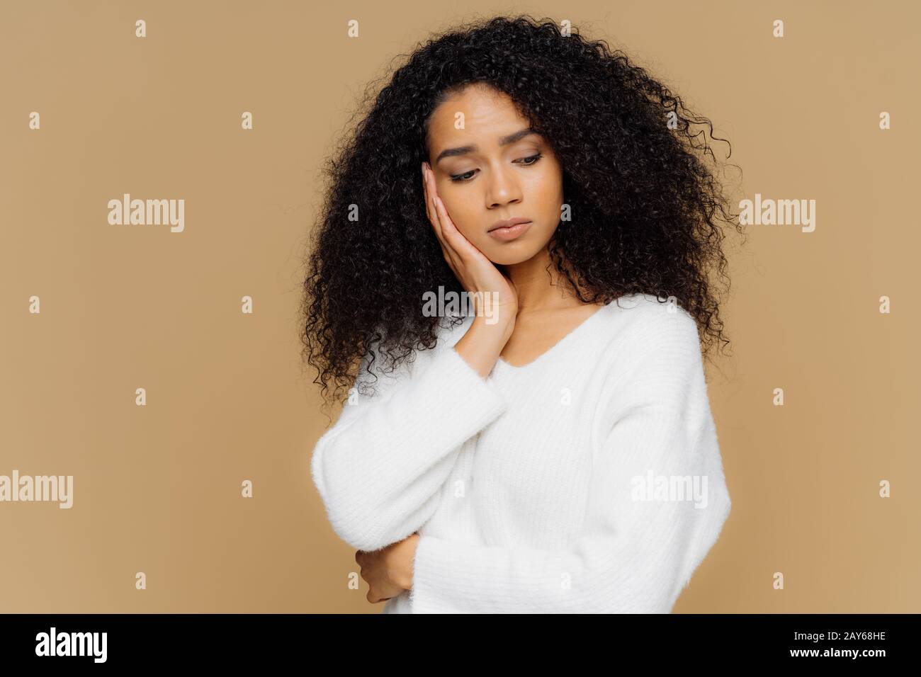 Melancholic sad lonely woman focused down with thoughtful expression, keeps hand on cheek, has calm expression, wears white sweater, has curly hair, i Stock Photo