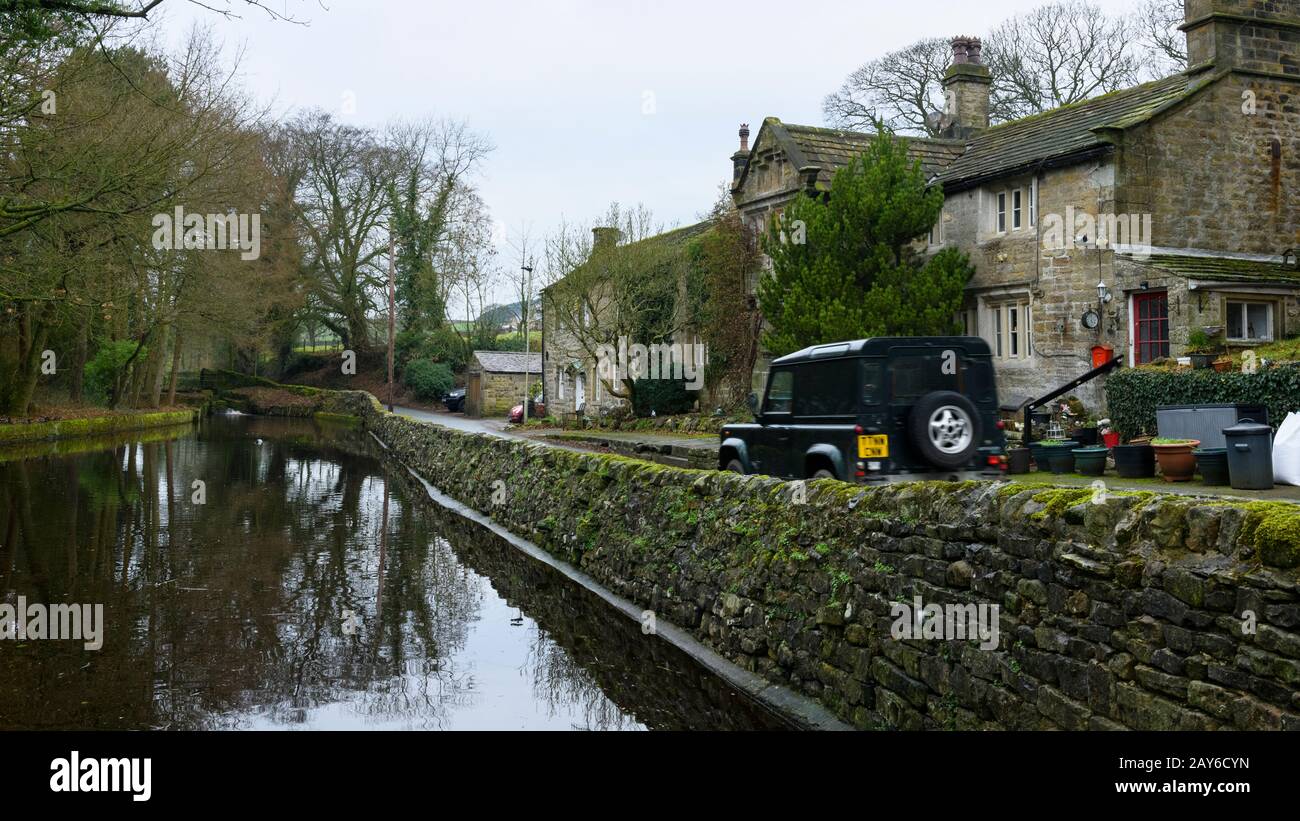 Land Rover Defender travelling & being driven on country road past quaint houses & historic mill pond - Embsay village, North Yorkshire, England, UK. Stock Photo