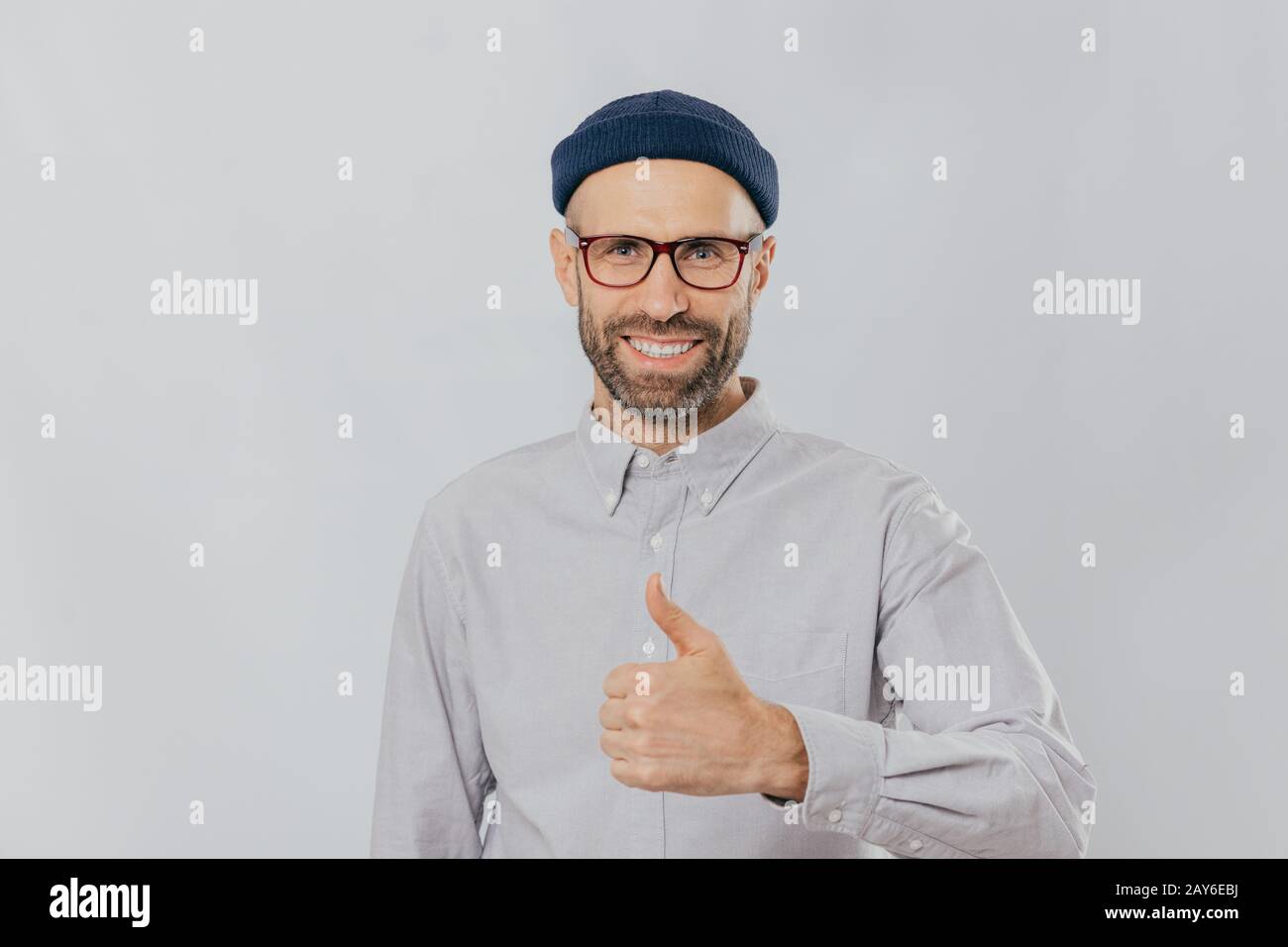 Positive smiling man with stubble, raises thumb up, demonstrates his like and approvement, wears headgear and formal shirt, isolated over white backgr Stock Photo