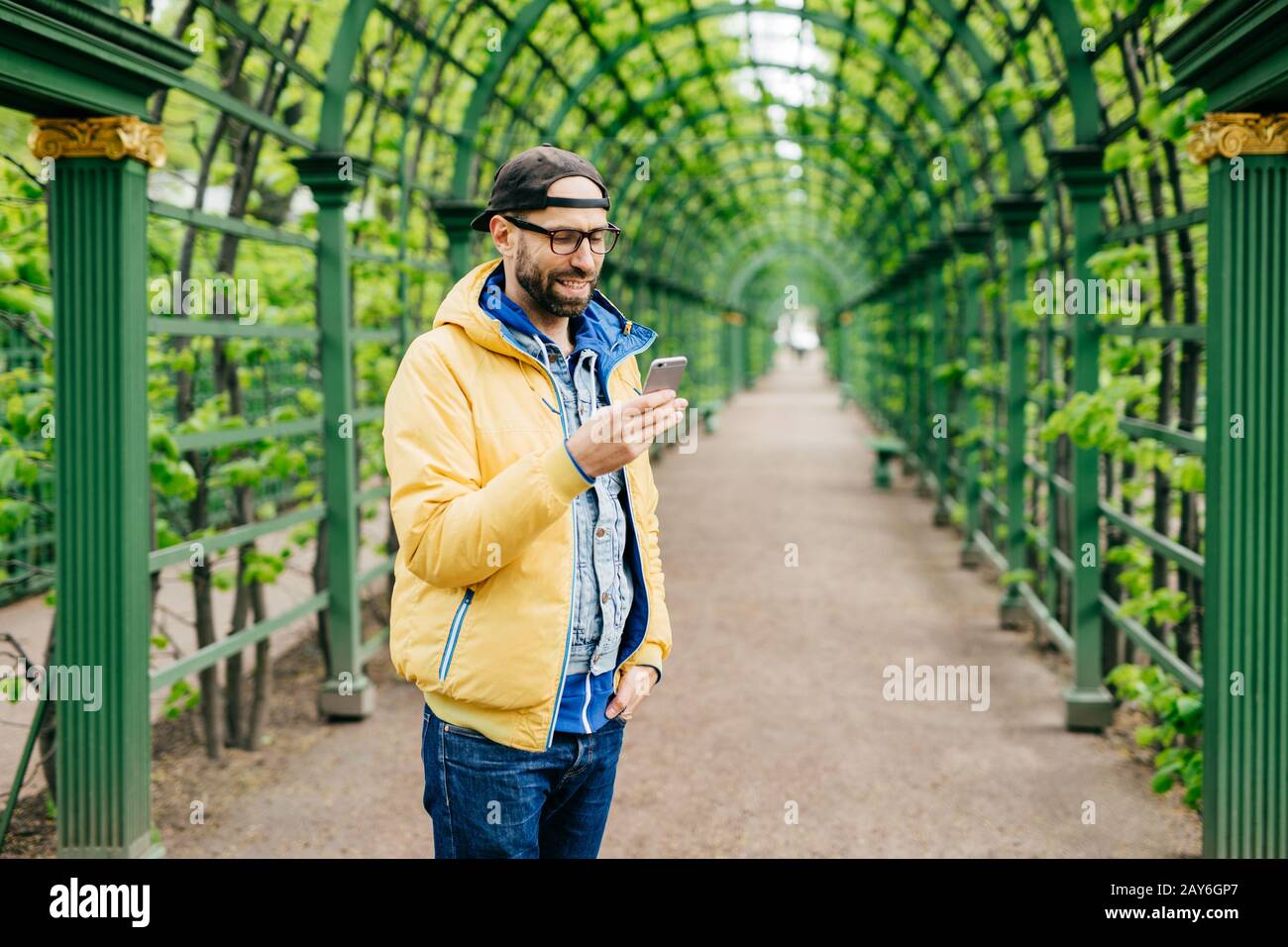 Trendy guy with beard and moustache wearing anorak, jeans and cap standing sideways having attentive look into his smartphone isolated over green arch Stock Photo