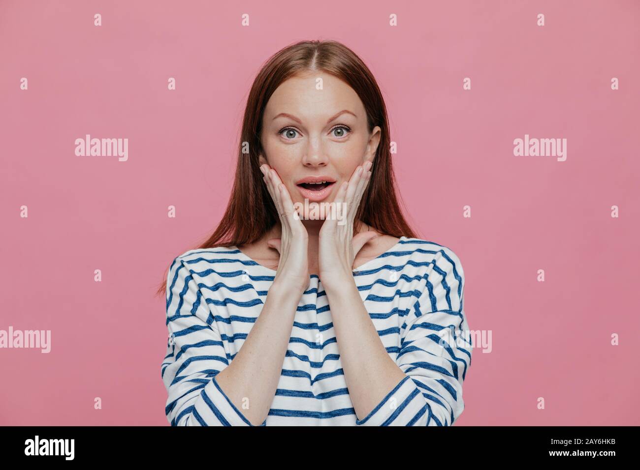 Beautiful dark haired amazed woman touches cheeks with both hands, dressed in striped clothes, looks surprisingly at camera, poses against pink backgr Stock Photo