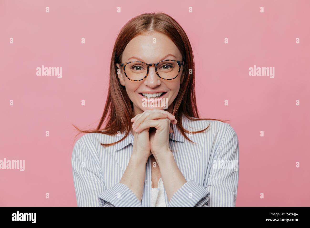 Positive young Caucasian woman keeps hands under chin, has dark hair, toothy smile, wears white shirt and glasses, models over pink background, listen Stock Photo