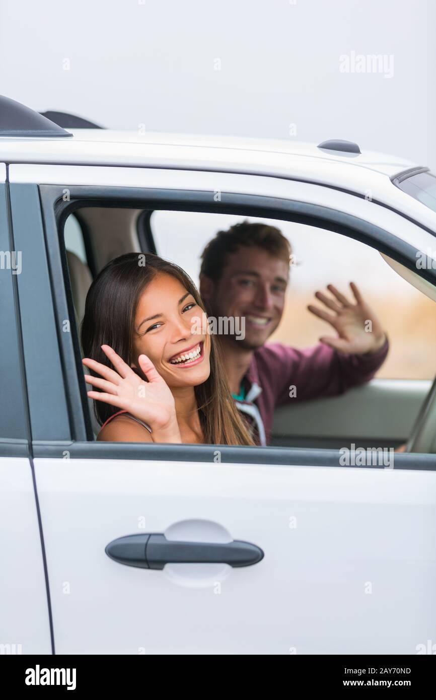 Happy multiracial couple leaving for road trip on summer vacation holiday. Young people driving car to travel destination waving goodbye. Stock Photo