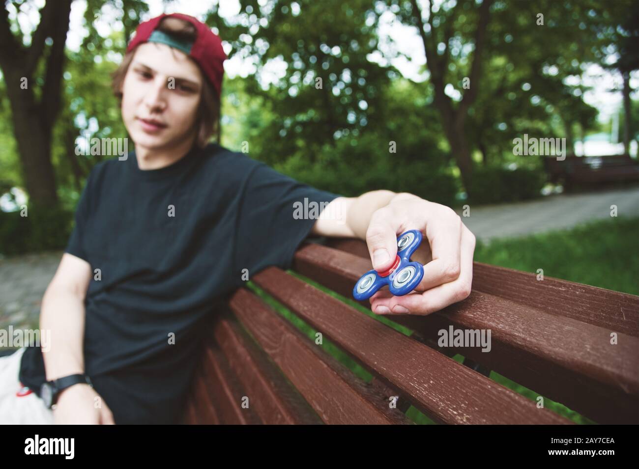 A long-haired hipster in a cap sits on a bench and spins a fidget-spinner Stock Photo