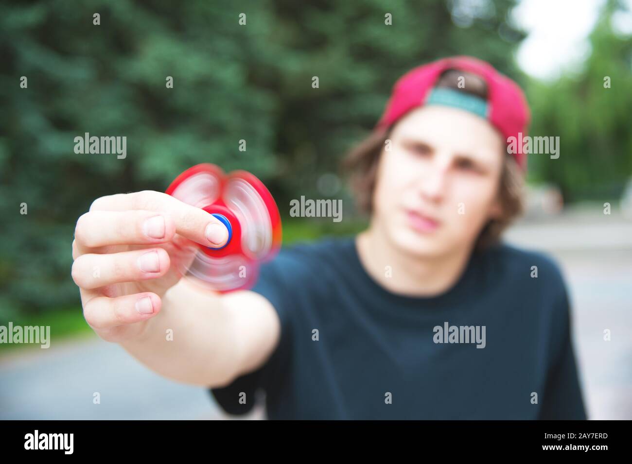 A long-haired hipster in a cap sits on a bench and spins a fidget-spinner Stock Photo