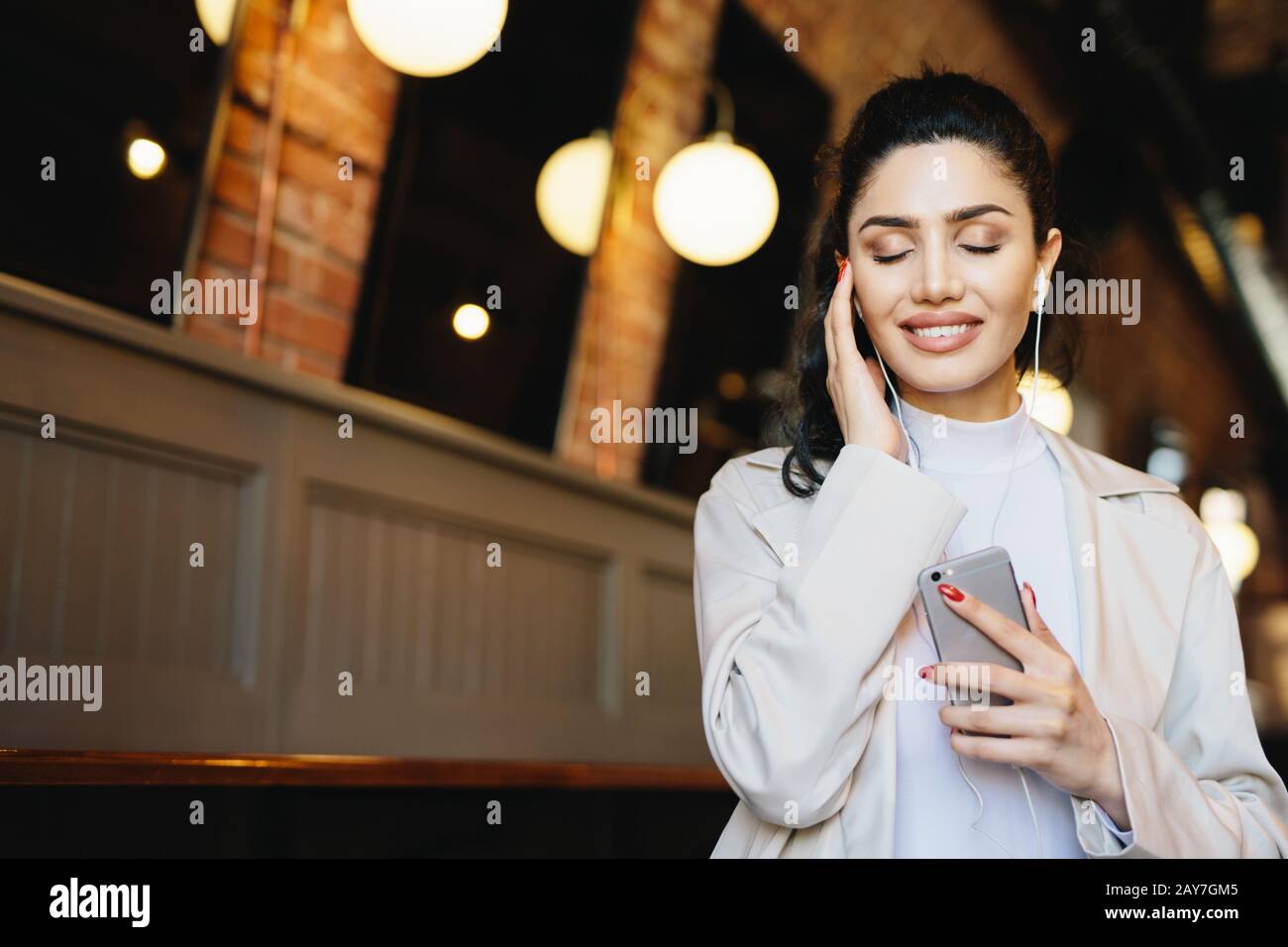 Restful brunette woman with lovely appearance closing her eyes with joy while enjoying loud music in her earphones. Stylish woman listening to her fav Stock Photo